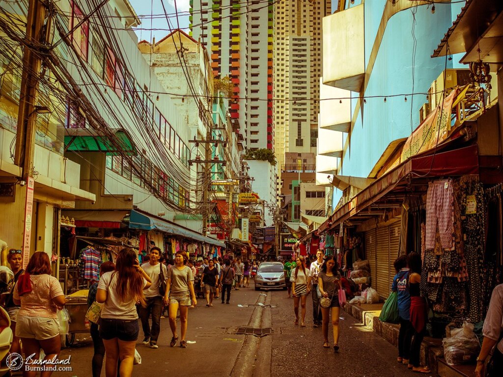 A street in Bangkok, Thailand, is filled with people, vehicles, and street vendors, as tall buildings tower over the scene. Read all about it at Burnsland!