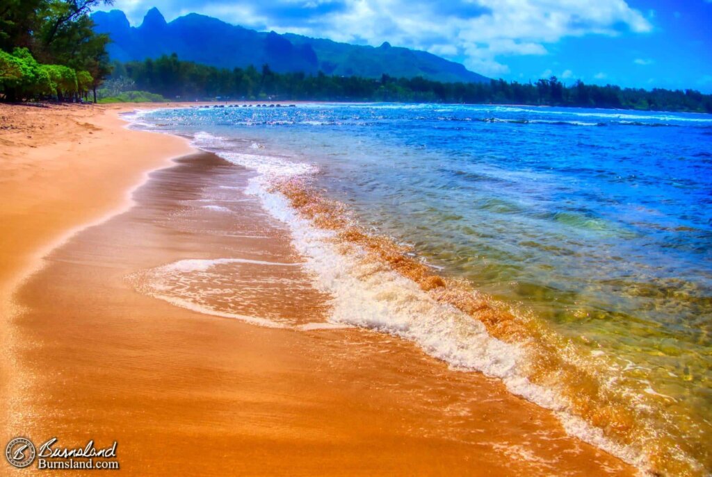 Waves roll onto the shore with blue water and mountains in the distance at Anahola Beach on the island of Kauaʻi in Hawaiʻi