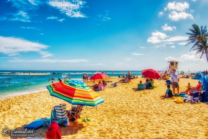 Beach goers and colorful umbrellas fill the sand under a beautiful blue sky at Poʻipū Beach Park on the island of Kauaʻi in Hawaiʻi